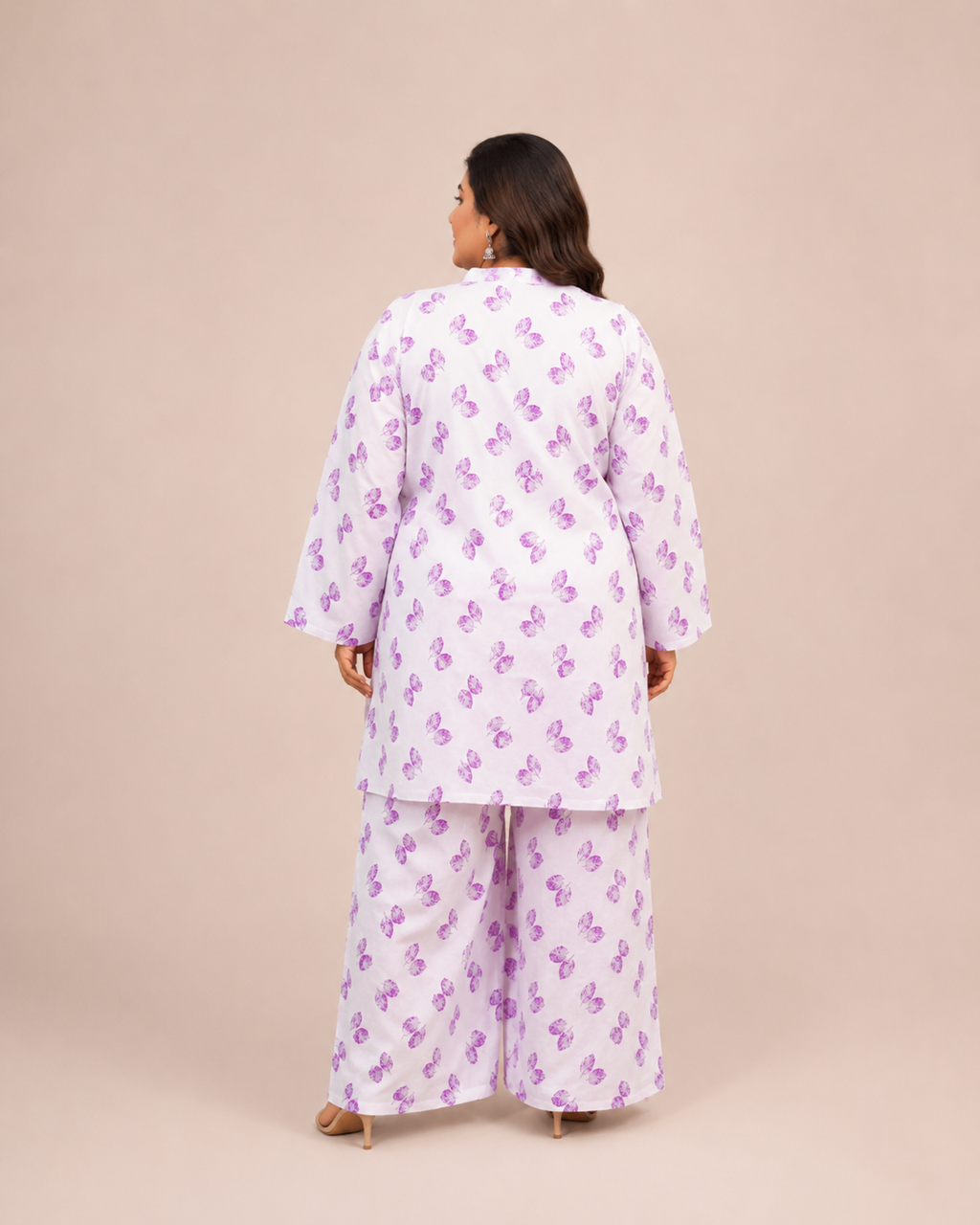 A woman viewed from the back wearing a matching pajama set with a white base and purple butterfly print, standing against a solid beige studio backdrop.